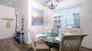 A dining area with a round glass table, beige chairs, and a view of a coastal building through large windows. - Beachside Cottage 2BR-3BA. Now booking for Summer! (Orange Beach)