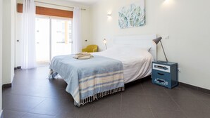 A hotel room with a bed, a chair, a desk with a lamp, a painting on the wall, and a door with curtains.