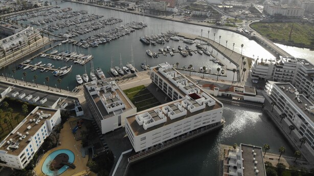 An aerial view of a marina with numerous boats docked, surrounded by modern buildings with flat roofs and a large swimming pool.