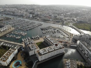 An aerial view of a marina with numerous boats docked, surrounded by modern buildings with flat roofs and a large swimming pool.
