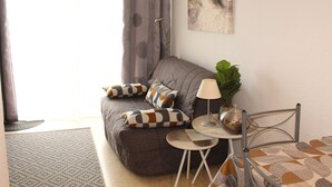 A living room with a grey sofa, patterned cushions, a small round table with a lamp, and a metal chair.