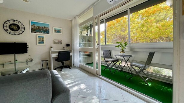 Una sala moderna con un sofá gris, un escritorio con una silla, un televisor y un ventanal con vistas a árboles otoñales.