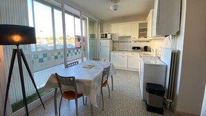 A compact kitchen with white cabinets, a dining table with chairs, a small refrigerator, and a window with a view of parked cars.
