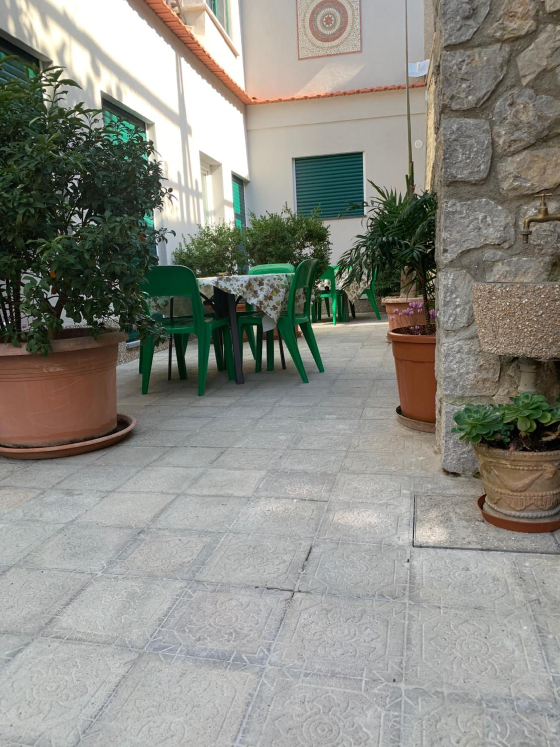 Un patio al aire libre con sillas y mesas verdes, rodeado de plantas en macetas y un muro de piedra.