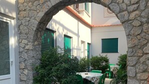 Ein Innenhof mit einem steinernen Torbogen, Blumentöpfen, einem Tisch mit Stühlen und einem Gebäude mit grünen Fensterläden.