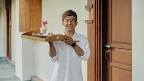 A person holding a tray with a flower and a plate of food, standing in front of a wooden door. - Paon Desa Ubud Hotel (Ubud)