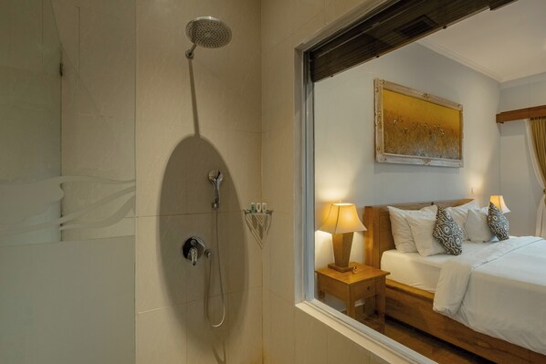 A hotel room with a shower, a bed with white bedding, a nightstand with a lamp, and a painting on the wall. - Paon Desa Ubud Hotel (Ubud)