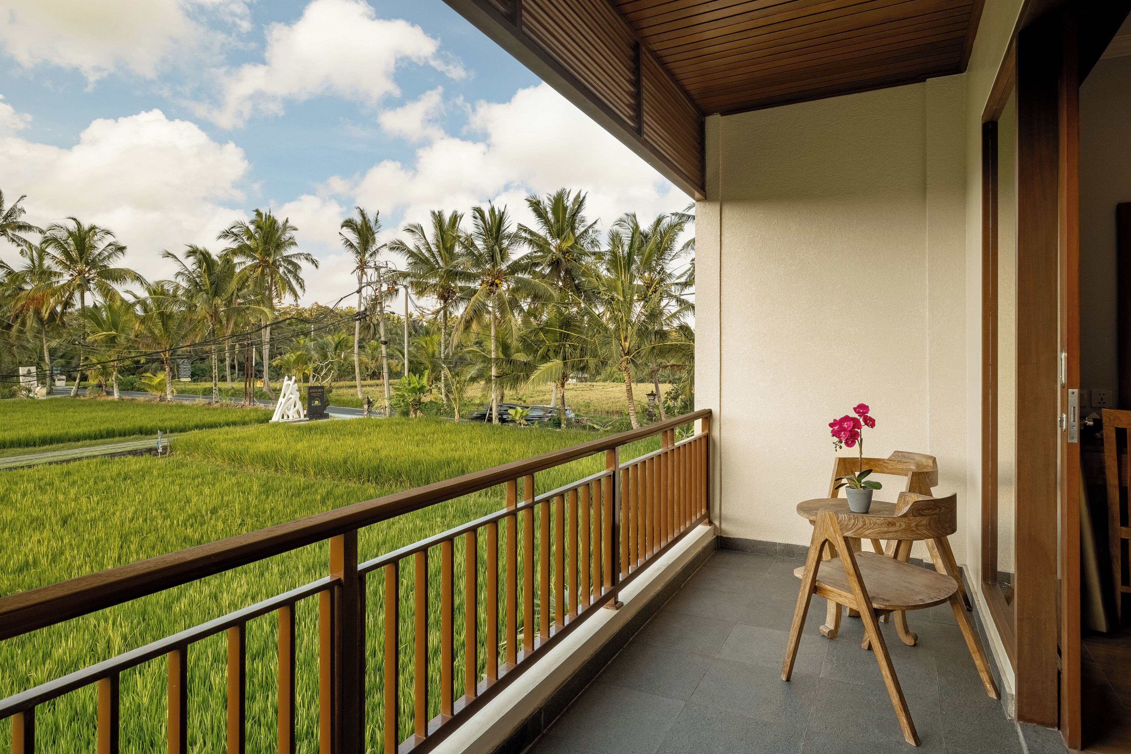 A balcony with a view of a green field and palm trees, a wooden chair, and a small table with a flower.