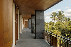 A covered corridor with stone pillars, wooden beams, and a balcony with a railing overlooking a tropical landscape. - Paon Desa Ubud Hotel (Ubud)