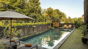 A swimming pool with lounge chairs and a stone wall, surrounded by lush greenery. - Paon Desa Ubud Hotel (Ubud)