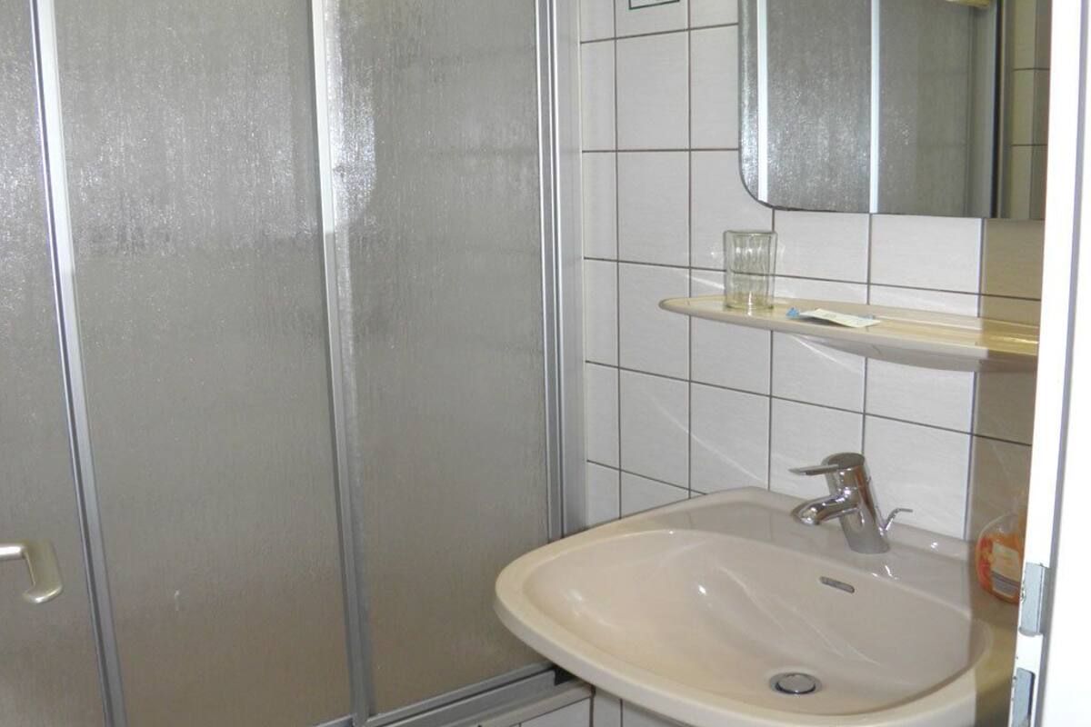 A bathroom with a shower, a sink, a mirror, and a shelf with a glass and a bottle.