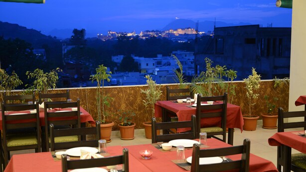 A rooftop restaurant with tables set for dining, potted plants, and a view of a cityscape at dusk. - Hotel Sanobar (Udaipur)