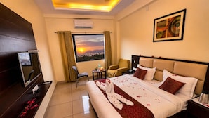 A hotel room with a bed, a desk with a computer, a chair, a small table with a bottle and flowers, and a window with a sunset view. - Hotel Sanobar (Udaipur)