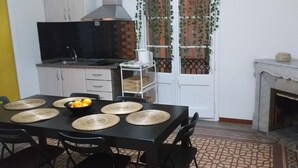 A compact kitchen with a gas stove, a sink, and a built-in range hood. There is a dining table with four placemats and a bowl of fruit. A small white shelf unit is against the wall, and a fireplace is visible in the corner. - V Barcelona Hostal (Barcelona)