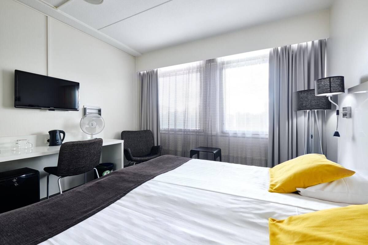 A hotel room with a bed, a desk, a chair, a TV, a window with curtains, and a lamp.