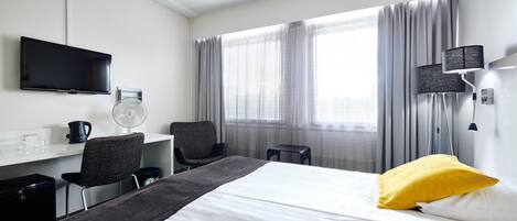 Una habitación de hotel con cama, escritorio, silla, televisor, ventana con cortinas y lámpara.