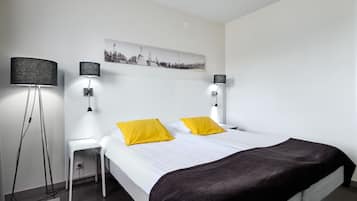 Una habitación de hotel moderna con una cama, dos lámparas de noche, una mesita de noche, una lámpara de pie y una obra de arte con un paisaje urbano en la pared.