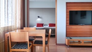 A hotel room with a wooden dining table, chairs, a TV mounted on the wall, and a bed with red pillows. - GreenStar Hotel Lahti (Lahti)
