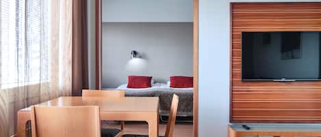 Habitación de hotel con una mesa de comedor de madera, sillas, un televisor montado en la pared y una cama con almohadas rojas.