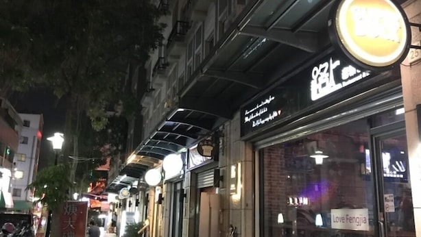 Cena noturna de rua com vitrines iluminadas, uma passarela para pedestres e motocicletas estacionadas.