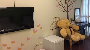 Quarto com televisão na parede, um urso de pelúcia em uma cadeira e um pequeno armário branco.