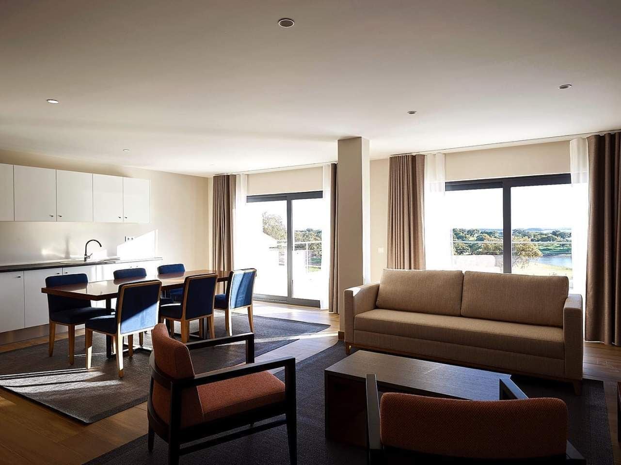 Una sala de estar moderna con sofá, mesa de centro y sillas, un comedor con mesa y sillas, y ventanas grandes con vistas.