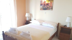 A neatly made bed with a white comforter and pillows, a wooden nightstand with a lamp, and a painting on the wall. - Corfu Island Apartment 129 (Corfu)