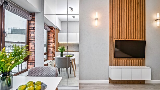 A modern living room with a flat-screen TV mounted on a wooden panel wall, a dining area with a white table and grey chairs, and a large window with a view of the city. - RentPlanet - Apartamenty Ruska (Wroclaw)