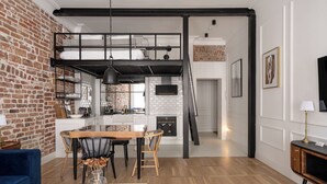 A modern loft-style apartment with exposed brick walls, a dining area with a wooden table and chairs, a kitchen with a black countertop, and a loft bedroom with a black metal staircase. - RentPlanet - Apartamenty Ruska (Wroclaw)