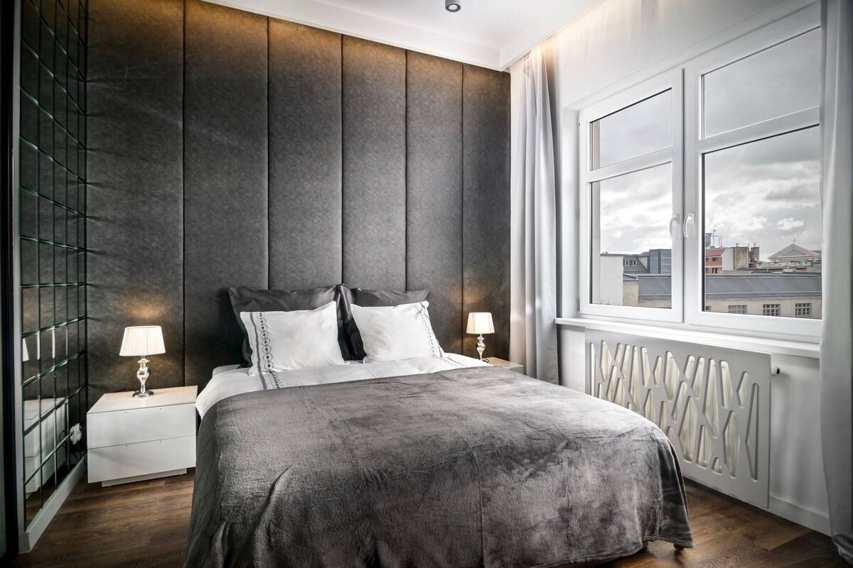 Une chambre moderne avec un grand lit, deux tables de chevet avec des lampes, une vue sur la ville par une fenêtre et un panneau décoratif au mur.