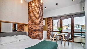 A modern hotel room with a large bed, a brick accent wall, a dining area with a table and chairs, and a view through large windows. - RentPlanet - Apartamenty Ruska (Wroclaw)