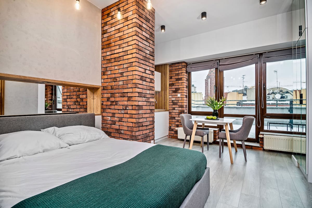 Une chambre d’hôtel moderne dotée d’un grand lit, d’un mur décoratif en briques, d’un coin repas avec une table et des chaises, et offrant une vue dégagée grâce à de grandes fenêtres.