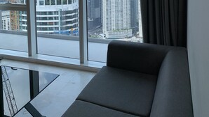 Una sala de estar moderna con un amplio ventanal que ofrece vistas a un paisaje urbano, incluyendo rascacielos y grúas de construcción.