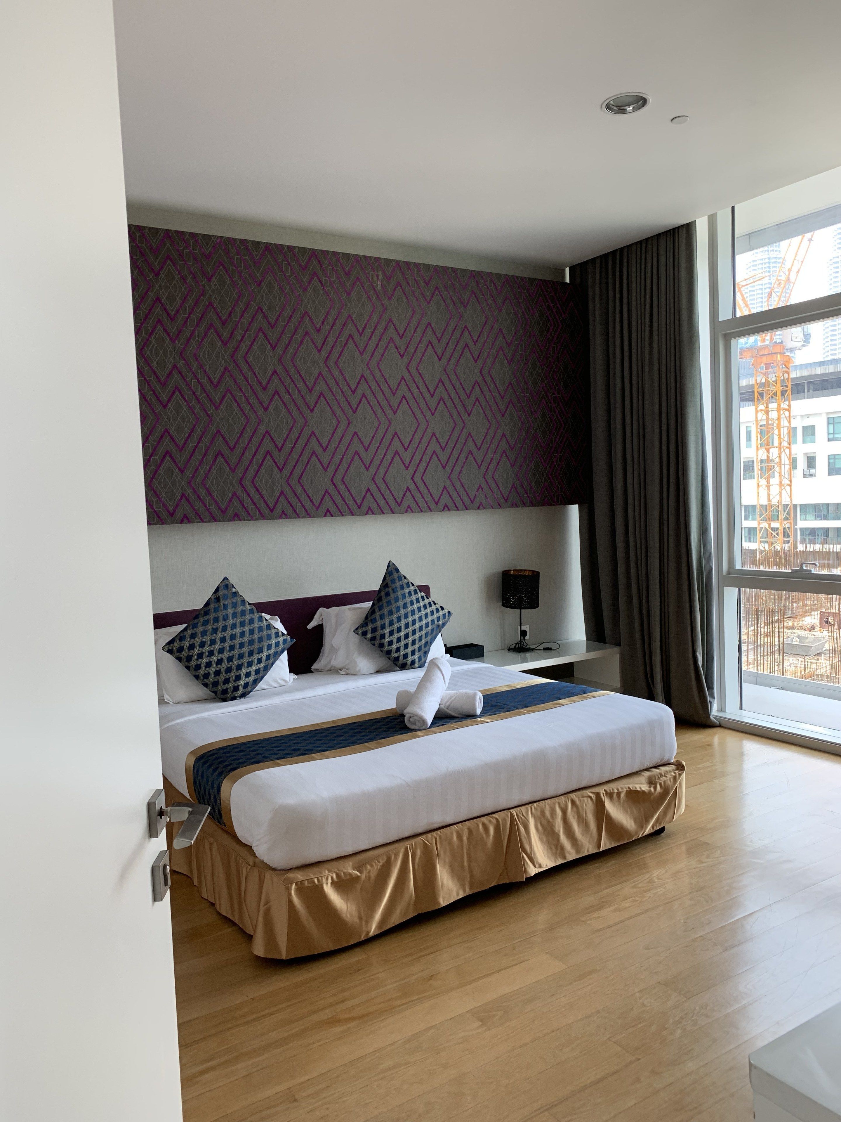 Ein Hotelzimmer mit einem großen Bett, einem Nachttisch, einer Lampe und Blick auf eine Baustelle durch das Fenster.