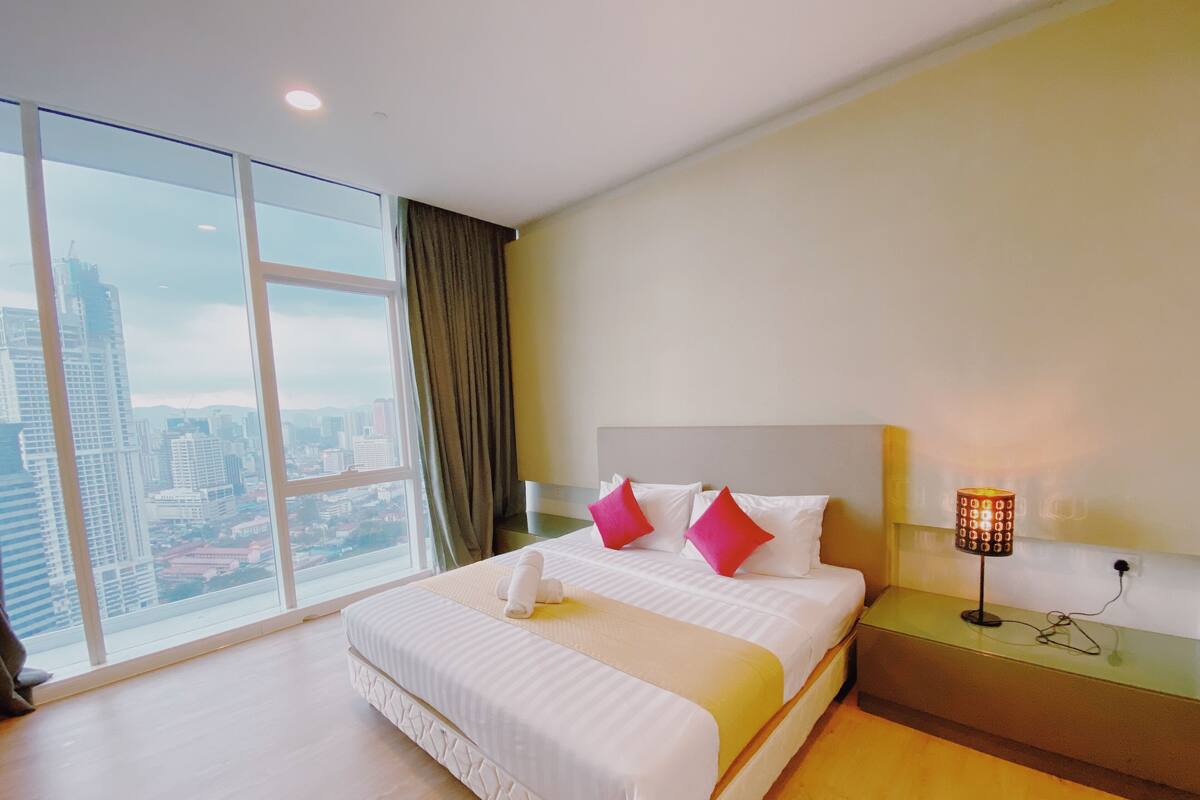 A hotel room with a large window, a bed with white bedding and pink pillows, a bedside table with a lamp, and a view of a cityscape.