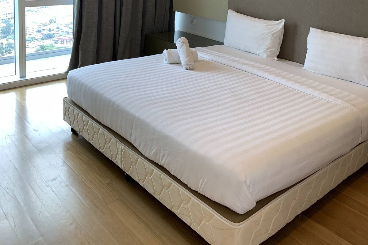 A hotel room with a large bed, a nightstand, a window with curtains, and a view of the city.