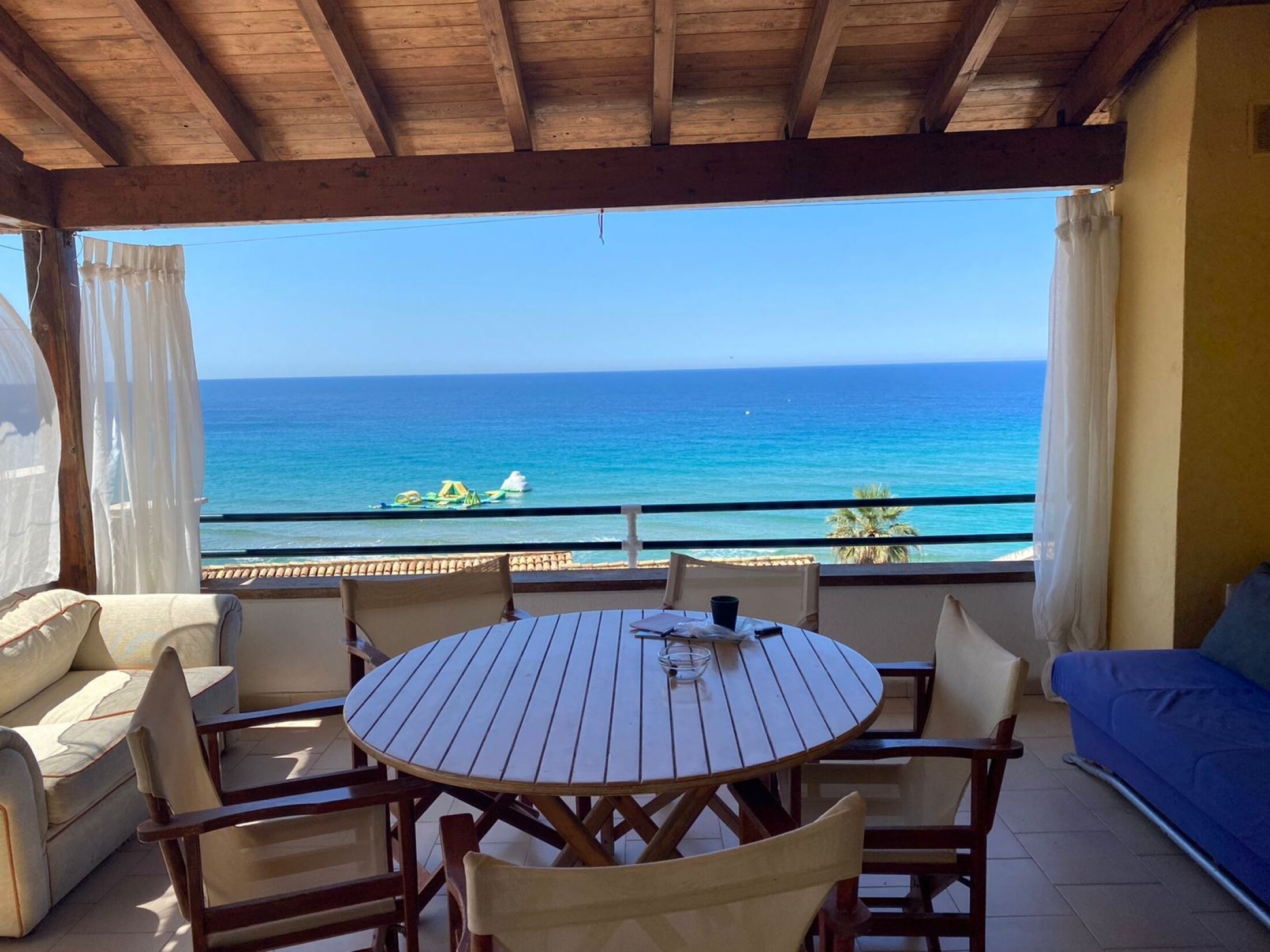 Un espace extérieur abrité, aménagé avec des sièges, offrant une vue sur la mer, comprenant une table ronde en bois et des chaises rembourrées.