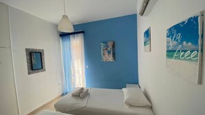 A room with two beds, a window with sheer curtains, a wall-mounted air conditioner, and a beach-themed poster. - Corfu Island Apartment 91 (Corfu)