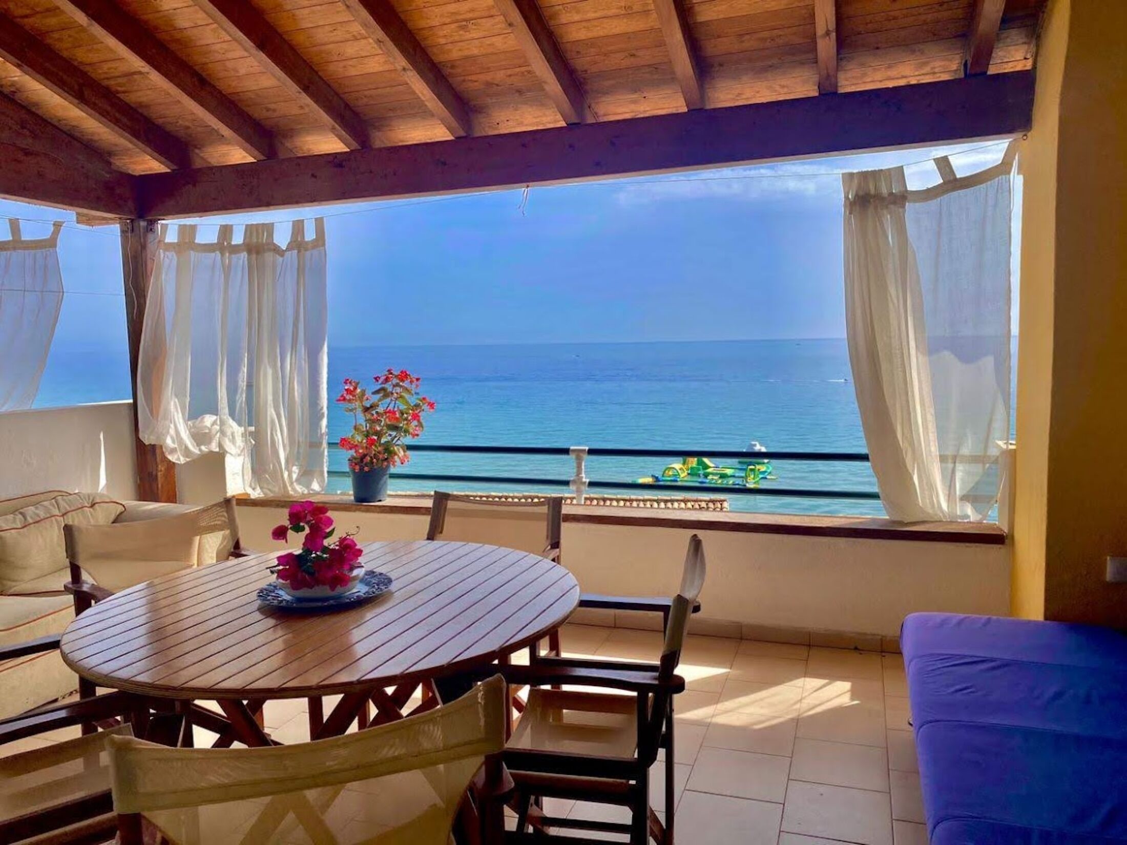 Uma área de estar coberta ao ar livre com mesa e cadeiras de madeira, um vaso de planta e vista para o mar.