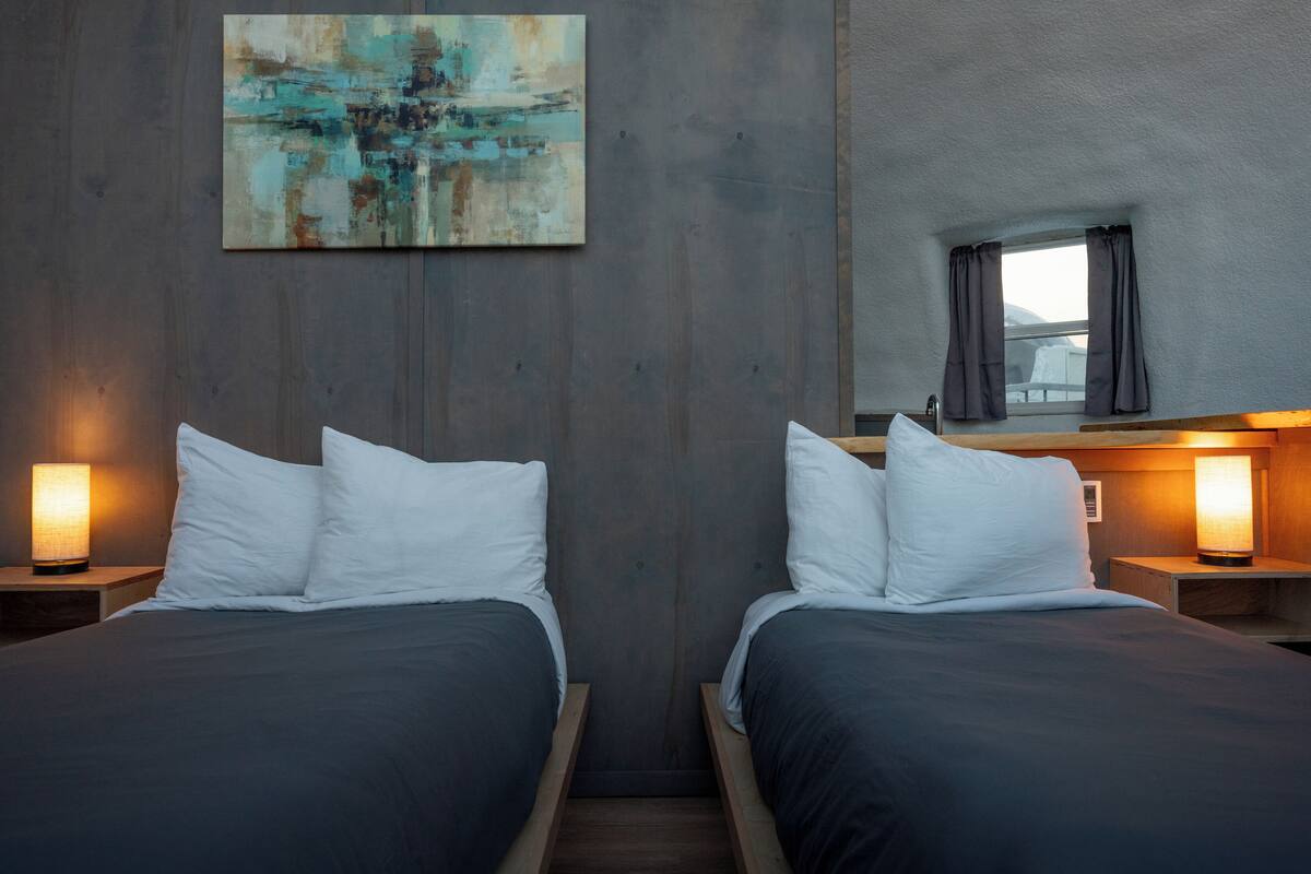 Habitación con dos camas, cada una con almohadas blancas y ropa de cama oscura, cabeceros de madera y lámparas de noche.