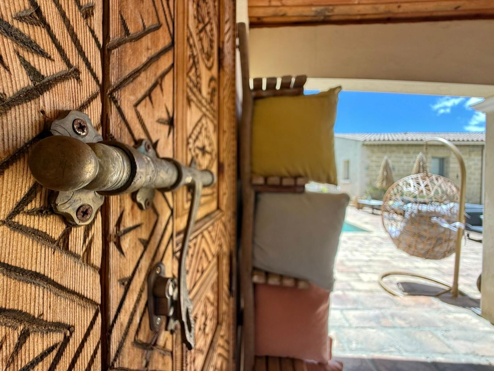 A wooden door with a metal latch and a window revealing a courtyard with a hanging chair and a building in the background.