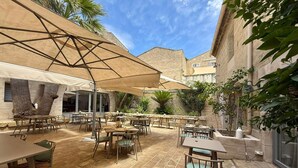 Une terrasse extérieure dotée de nombreuses tables et chaises, protégée par de grands parasols, adossée à un mur de pierre et entourée de verdure.