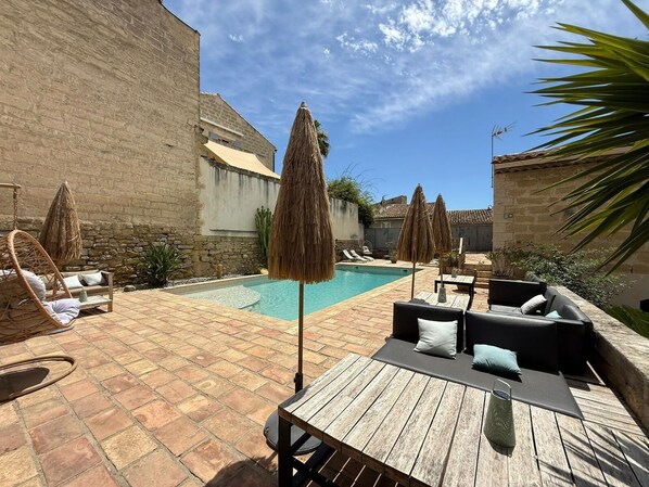 A pool area with lounge chairs, a wooden table, and thatched umbrellas. - Hôtel Restaurant O'Banel (Gallargues-le-Montueux)