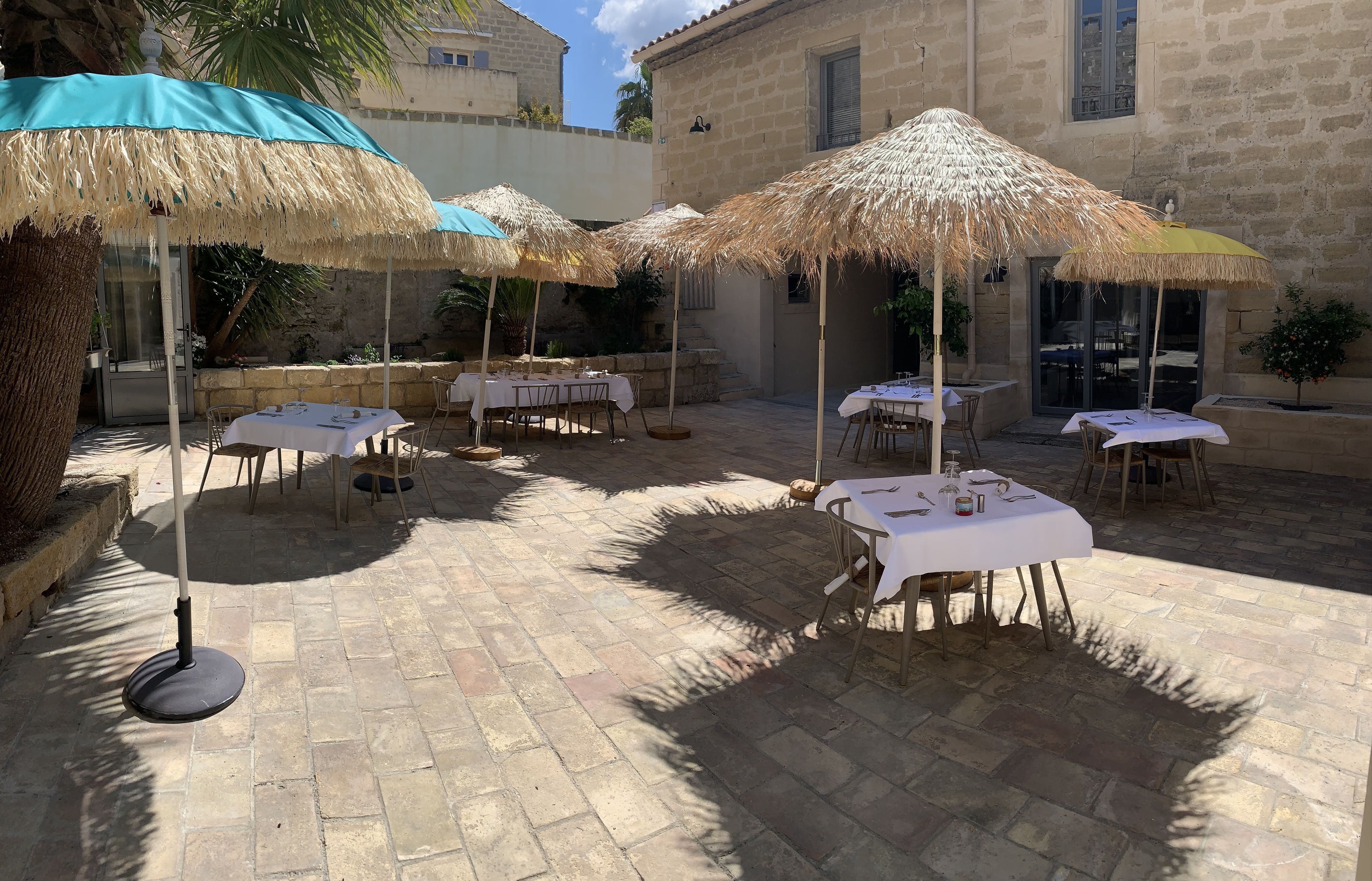 An outdoor patio with tables and thatched umbrellas, stone walls, and a palm tree.