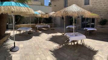An outdoor patio with tables and thatched umbrellas, stone walls, and a palm tree.