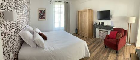 A hotel room with a large bed, a wooden wardrobe, a TV mounted on the wall, a red armchair, and a floor lamp.