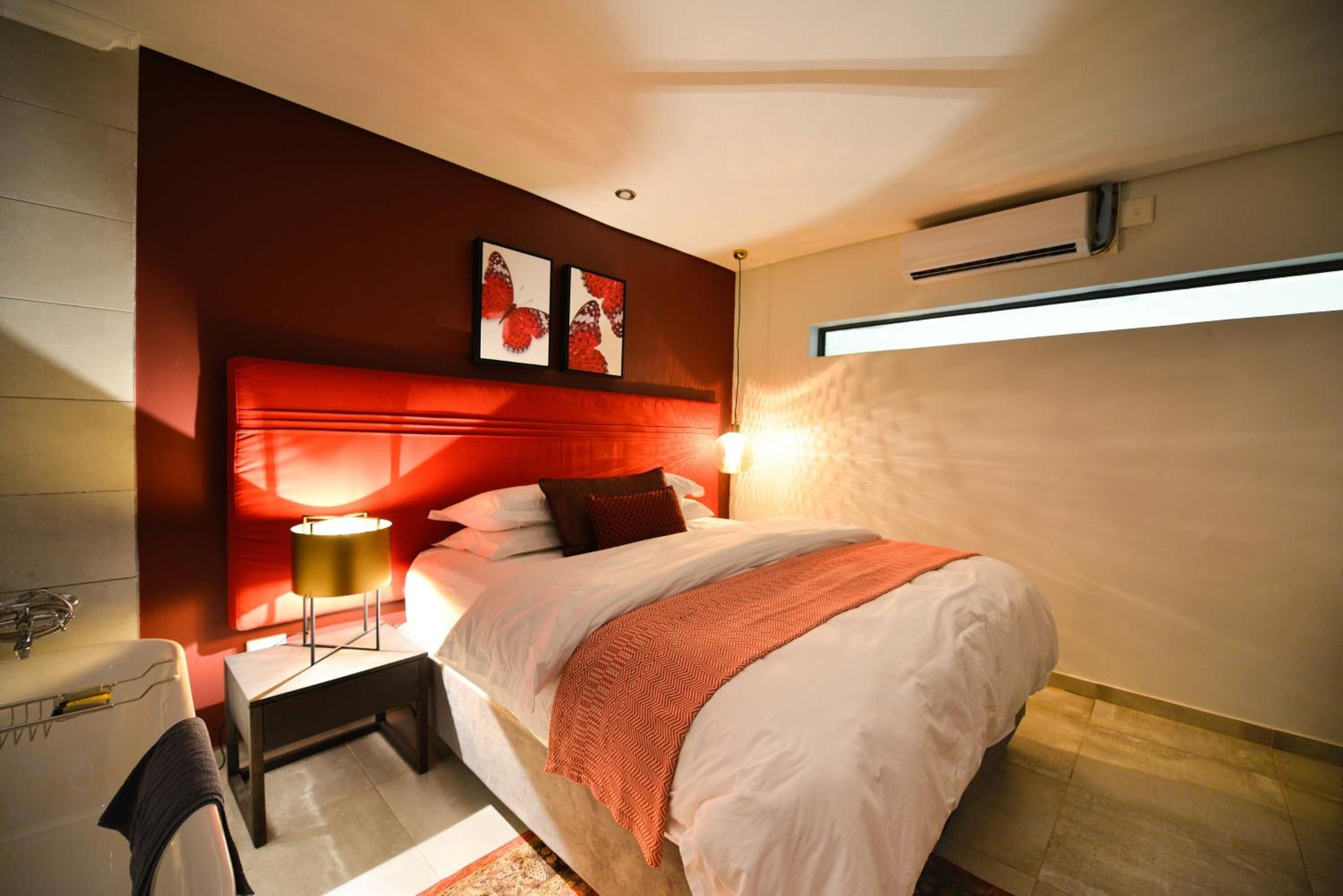 A modern bedroom with a red accent wall, a bed with white linens, a desk with a lamp, and a window with a view.