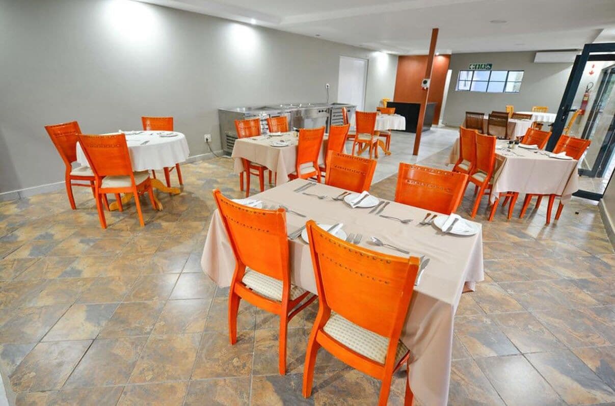 Un comedor con sillas naranjas y manteles blancos, un área de cocina y vista al exterior a través de un ventanal.