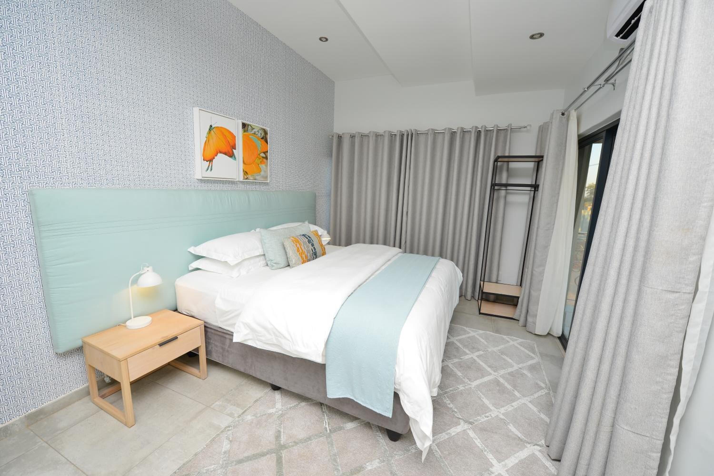 A bedroom with a bed, a nightstand, a window with curtains, and a wall with a geometric pattern.
