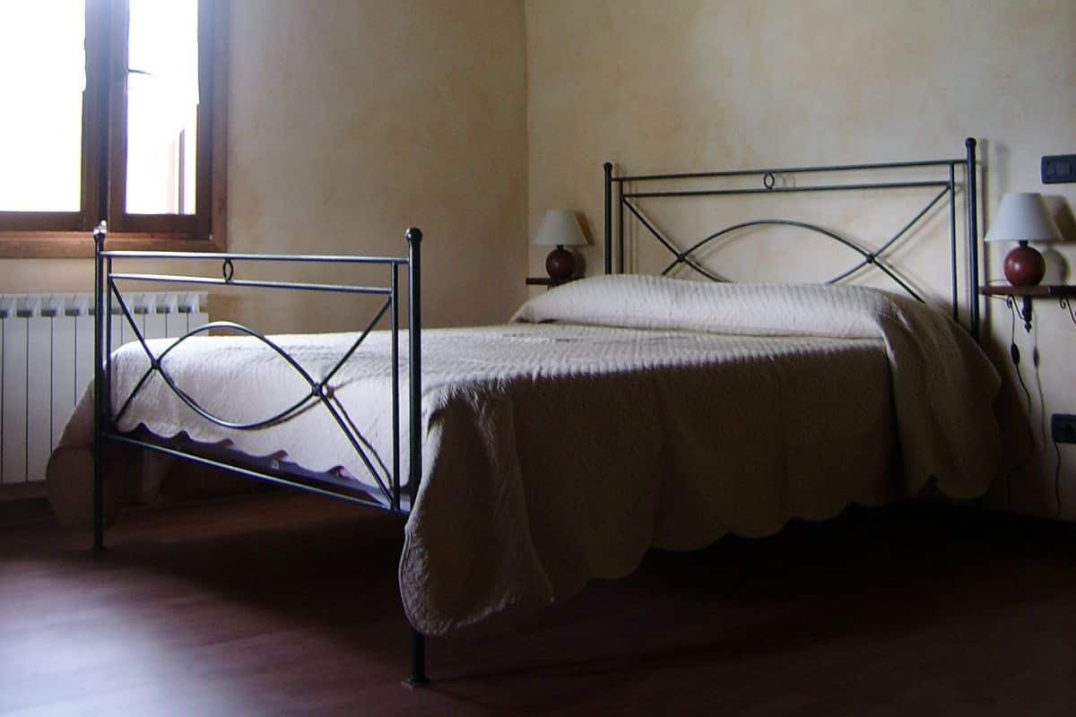 A room with two metal-framed beds, a window, a radiator, and a wall-mounted lamp.